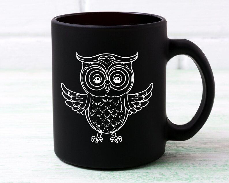 May include: Black ceramic coffee mug with a white outline of an owl with large eyes. The owl is facing forward with its wings spread out.