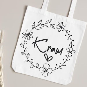 Pu&ograve; includere: Borsa tote bianca con un design a ghirlanda floreale nera, la parola "Kraw" e un simbolo a cuore. La borsa ha due manici ed &egrave; realizzata in tessuto bianco semplice. Il design &egrave; centrato.