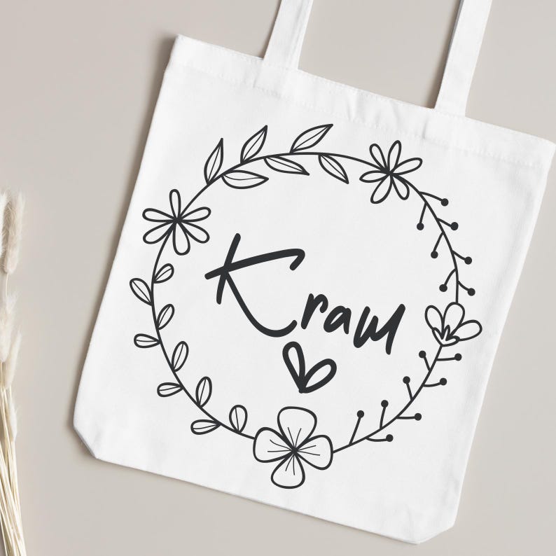 Pu&ograve; includere: Borsa tote bianca con un design a ghirlanda floreale nera, la parola "Kraw" e un simbolo a cuore. La borsa ha due manici ed &egrave; realizzata in tessuto bianco semplice. Il design &egrave; centrato.