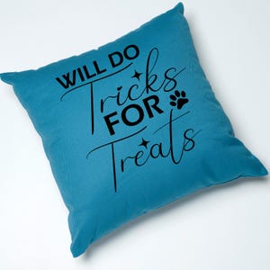 Puede incluir: Una almohada de color azul turquesa con el texto "Will Do Tricks For Treats" en letras negras. Tambi&eacute;n se incluye una huella de pata y dos estrellas en el dise&ntilde;o.