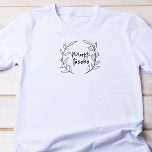 Pu&ograve; includere: T-shirt bianco con scollo tondo. La maglietta presenta un motivo a ghirlanda floreale nera con le parole "Markt-Tasche" al centro. Il disegno &egrave; centrato sul petto della maglietta.