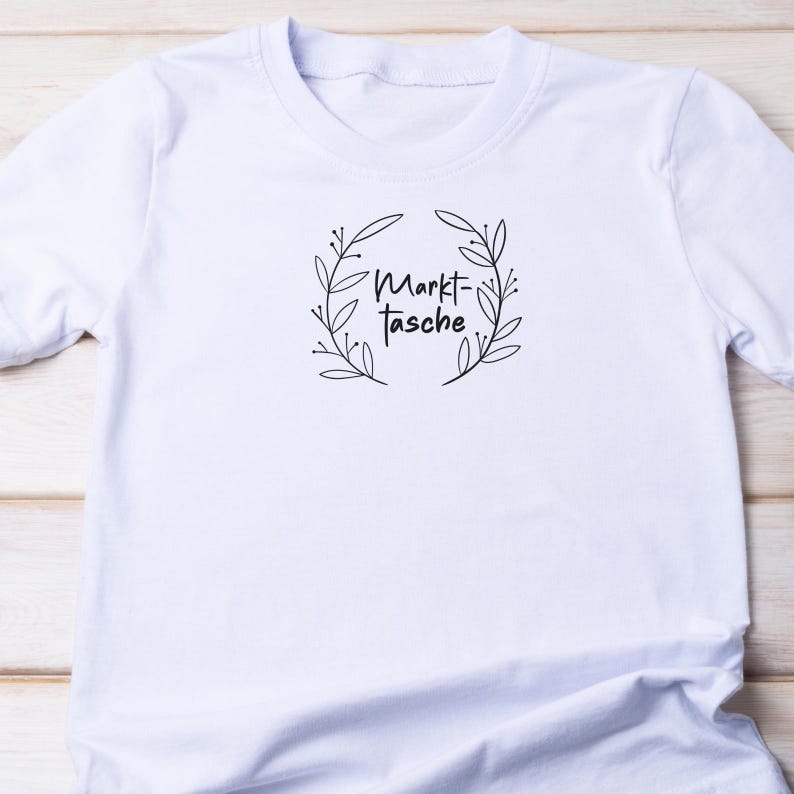 Pu&ograve; includere: T-shirt bianco con scollo tondo. La maglietta presenta un motivo a ghirlanda floreale nera con le parole "Markt-Tasche" al centro. Il disegno &egrave; centrato sul petto della maglietta.