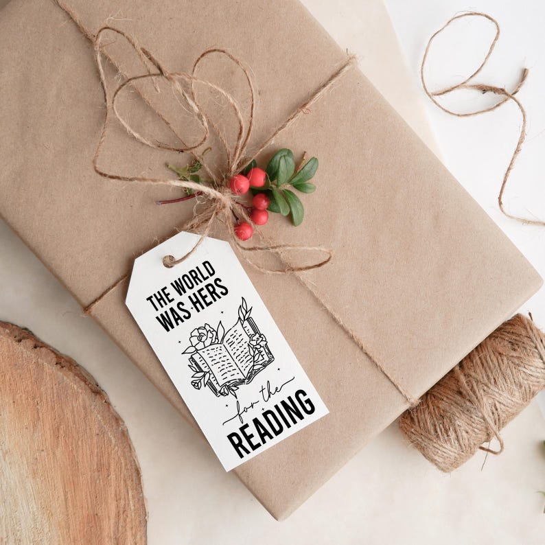 May include: A white gift tag with black text and a black and white illustration of an open book with flowers. The text reads "The world was hers for the reading."