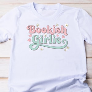 K&ouml;nnte beinhalten: Wei&szlig;es T-Shirt mit dem Aufdruck "Bookish Girlie" in Rosa und Mintgr&uuml;n, verziert mit Blumen und Herzen. Das Design ist auf einem leichten, weichen Stoff gedruckt, ideal f&uuml;r den t&auml;glichen Gebrauch.