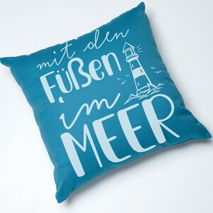Puede incluir: Una almohada de color azul turquesa con texto blanco que dice "mit den F&uuml;&szlig;en im MEER" y una silueta blanca de un faro con olas y p&aacute;jaros.