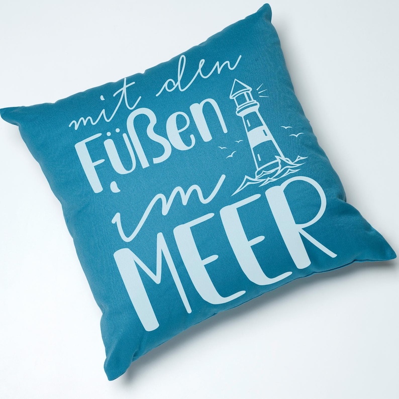 Puede incluir: Una almohada de color azul turquesa con texto blanco que dice "mit den F&uuml;&szlig;en im MEER" y una silueta blanca de un faro con olas y p&aacute;jaros.