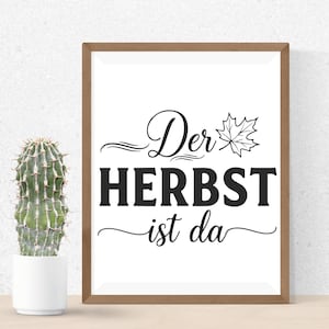 May include: A framed print with the words "Der HERBST ist da" in black script and block letters, with a maple leaf illustration. The frame is brown, and a cactus in a white pot is to the left.