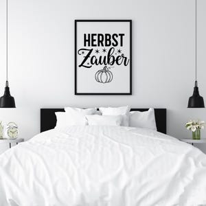 May include: A framed black and white print with the words "HERBST Zauber" and a pumpkin illustration. The artwork is displayed above a bed with white bedding. The room has a minimalist design with black pendant lights.