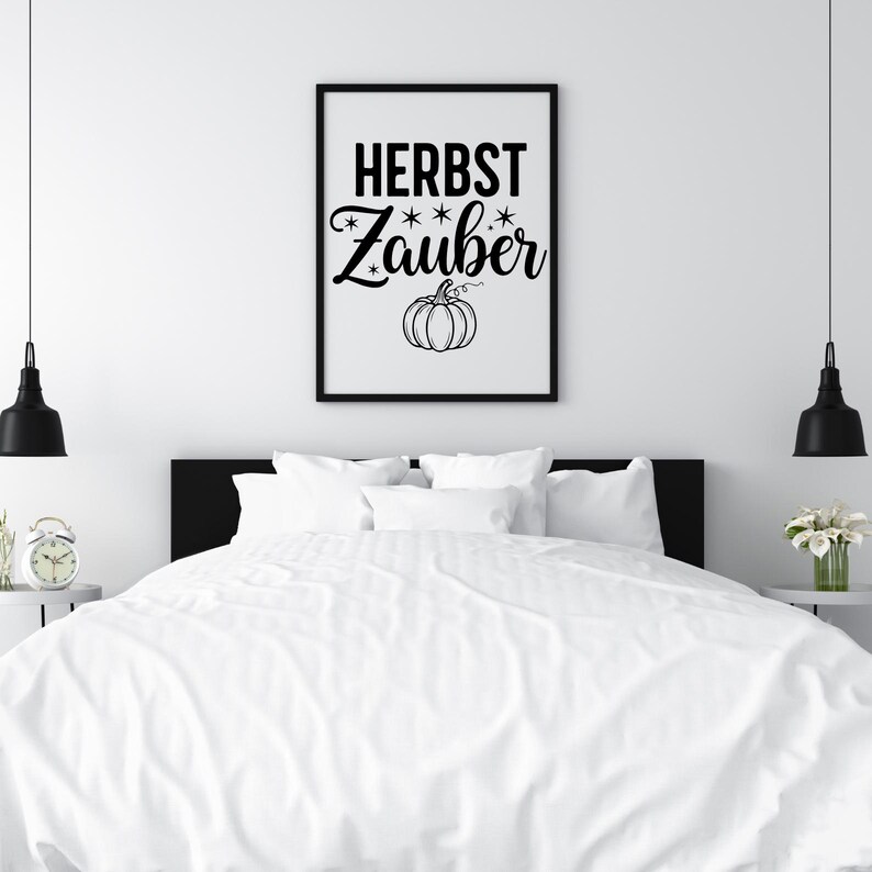 May include: A framed black and white print with the words "HERBST Zauber" and a pumpkin illustration. The artwork is displayed above a bed with white bedding. The room has a minimalist design with black pendant lights.