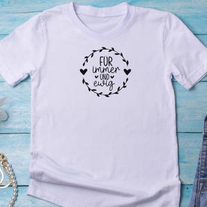 May include: White t-shirt with a black graphic that says "F&uuml;r immer und ewig" with a heart on either side of "und" and a wreath around the text.