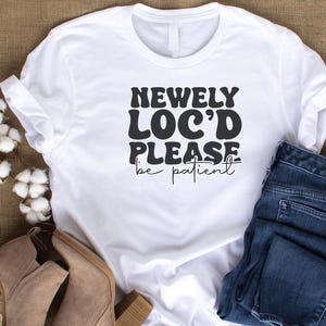May include: White t-shirt with black text that reads "Newly Loc'd Please be patient". The shirt is laying on a brown surface with a pair of blue jeans and brown boots.