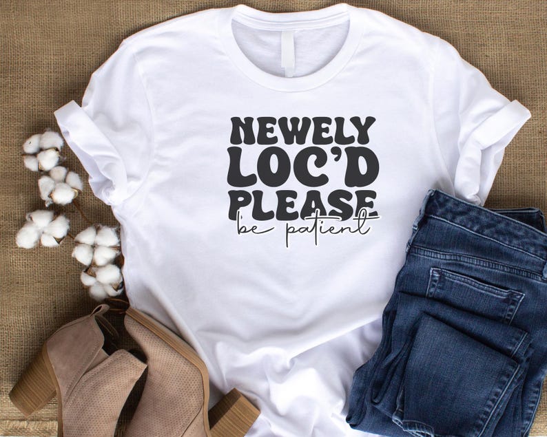May include: White t-shirt with black text that reads "Newly Loc'd Please be patient". The shirt is laying on a brown surface with a pair of blue jeans and brown boots.