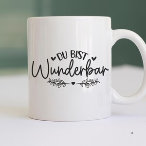 May include: White ceramic mug with black text that reads "Du bist wunderbar" with a heart and leafy branches on either side of the text.