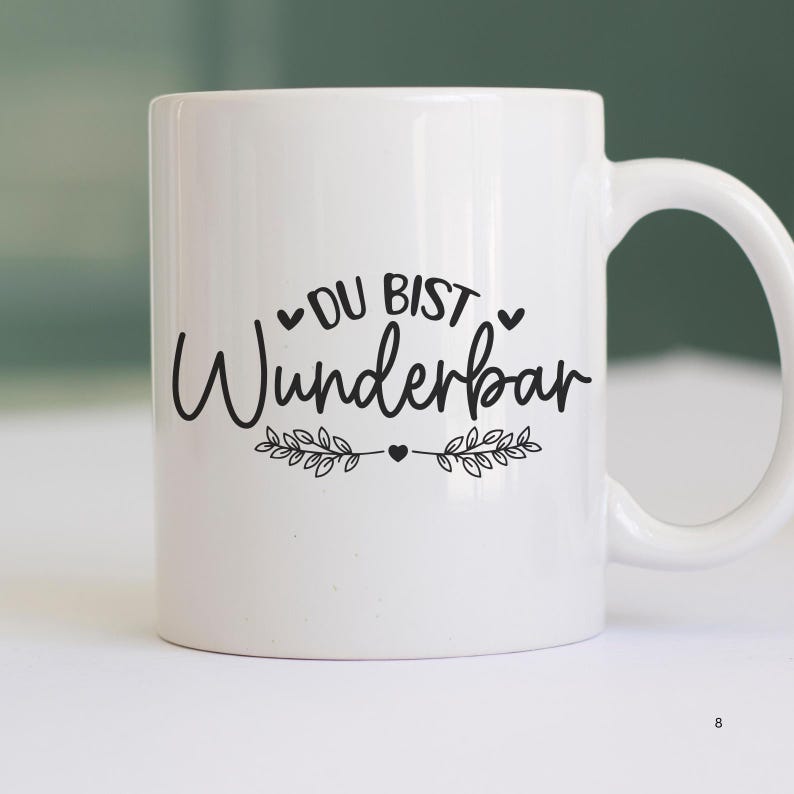 May include: White ceramic mug with black text that reads "Du bist wunderbar" with a heart and leafy branches on either side of the text.
