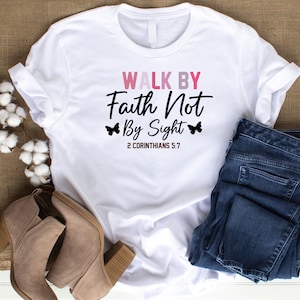 May include: White t-shirt with the text "Walk by Faith Not By Sight" and the scripture reference "2 Corinthians 5:7". The text is in black with pink and purple accents. There are two butterfly graphics on either side of the text.