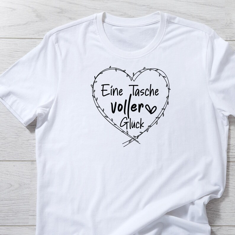 Pu&ograve; includere: T-shirt bianca con un disegno nero a forma di cuore. Il cuore contiene il testo tedesco "Eine Tasche voller Gl&uuml;ck", che si traduce in "Una borsa piena di felicit&agrave;". Il design &egrave; semplice ed elegante.