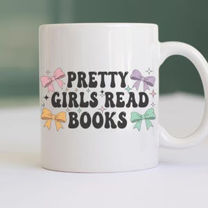 K&ouml;nnte beinhalten: Wei&szlig;er Keramikbecher mit dem Text "PRETTY GIRLS READ BOOKS" in Schwarz, verziert mit bunten Schleifen. Der Becher hat einen gebogenen Henkel und ist f&uuml;r Getr&auml;nke konzipiert.