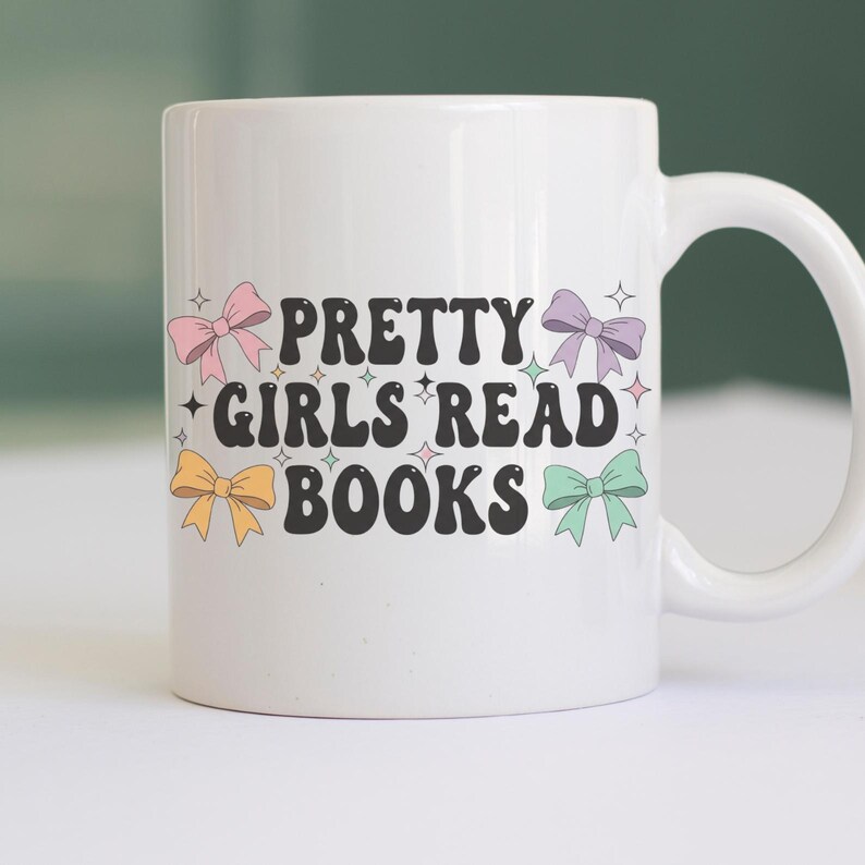 K&ouml;nnte beinhalten: Wei&szlig;er Keramikbecher mit dem Text "PRETTY GIRLS READ BOOKS" in Schwarz, verziert mit bunten Schleifen. Der Becher hat einen gebogenen Henkel und ist f&uuml;r Getr&auml;nke konzipiert.