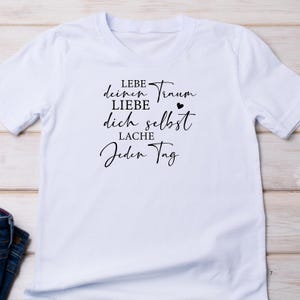 May include: White t-shirt with black text that reads "Lebe deinen Traum LIEBE dich selbst LACHE Jeden Tag".
