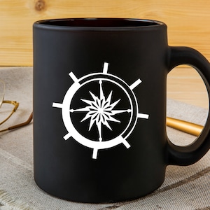 May include: A black ceramic mug with a white compass design. The compass has a star in the center and is surrounded by a circle.