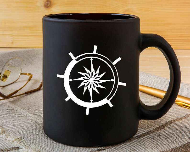 May include: A black ceramic mug with a white compass design. The compass has a star in the center and is surrounded by a circle.
