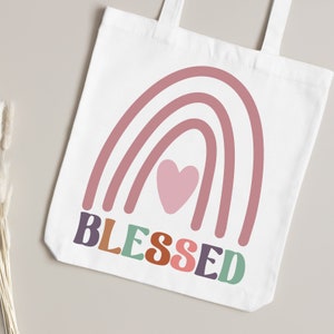 May include: A white tote bag with a pink rainbow design and a pink heart in the center. The word "BLESSED" is written in colorful letters below the rainbow.