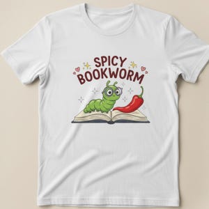 K&ouml;nnte beinhalten: Wei&szlig;es T-Shirt mit einem gr&uuml;nen Cartoon-B&uuml;cherwurm mit Brille, der auf einem offenen Buch neben einer roten Chilischote sitzt. Der Text "Spicy Bookworm" steht in Burgunderrot &uuml;ber dem B&uuml;cherwurm.