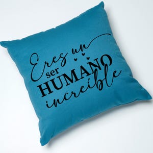 May include: A teal blue throw pillow with the text "Eres un ser HUMANO incre&iacute;ble" in black lettering. The text is written in a cursive font and includes three small hearts.