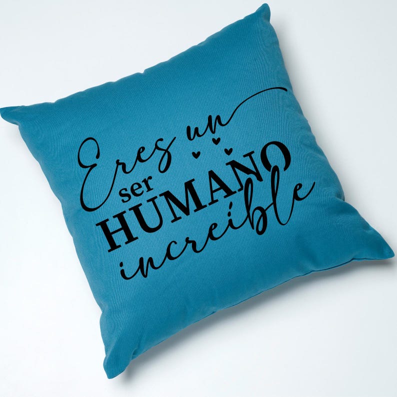 May include: A teal blue throw pillow with the text "Eres un ser HUMANO incre&iacute;ble" in black lettering. The text is written in a cursive font and includes three small hearts.