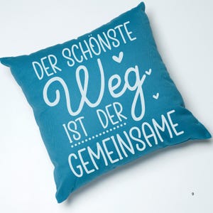 May include: A teal blue throw pillow with white text that reads "Der sch&ouml;nste Weg ist der gemeinsame" which translates to "The most beautiful path is the shared one".