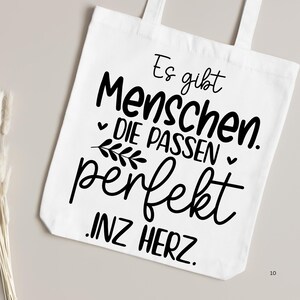 May include: White tote bag with black text that reads "Es gibt Menschen. Die passen perfekt. Inz Herz."