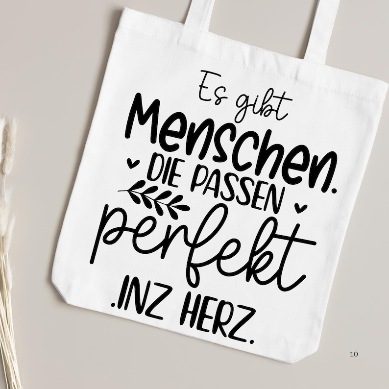 May include: White tote bag with black text that reads "Es gibt Menschen. Die passen perfekt. Inz Herz."
