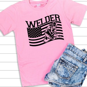 Puede incluir: Camiseta rosa con un gr&aacute;fico en blanco y negro de un soldador frente a una bandera estadounidense. El texto "Welder" est&aacute; sobre el gr&aacute;fico.