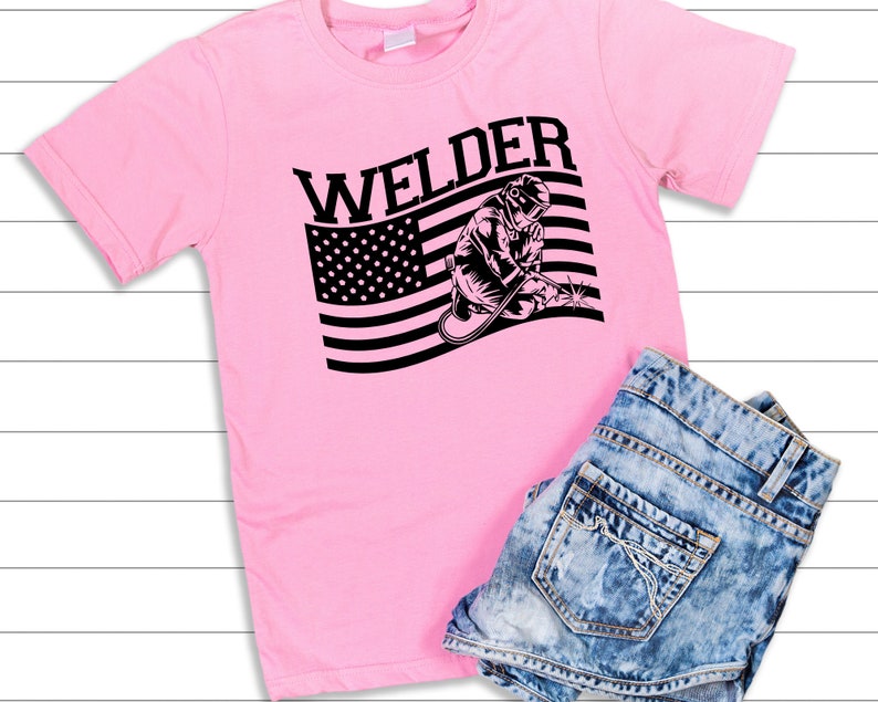 Puede incluir: Camiseta rosa con un gr&aacute;fico en blanco y negro de un soldador frente a una bandera estadounidense. El texto "Welder" est&aacute; sobre el gr&aacute;fico.