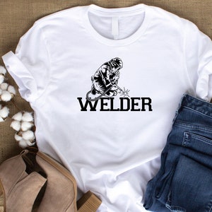Puede incluir: Camiseta blanca con una ilustraci&oacute;n en blanco y negro de un soldador y la palabra "WELDER" en texto negro.
