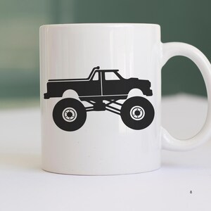 May include: White ceramic coffee mug with a black silhouette of a monster truck. The truck has large tires and a raised suspension.