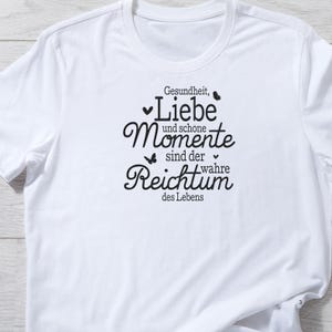 May include: White t-shirt with a black graphic print that reads "Gesundheit, Liebe und sch&ouml;ne Momente sind der wahre Reichtum des Lebens".