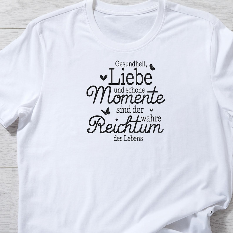May include: White t-shirt with a black graphic print that reads "Gesundheit, Liebe und sch&ouml;ne Momente sind der wahre Reichtum des Lebens".