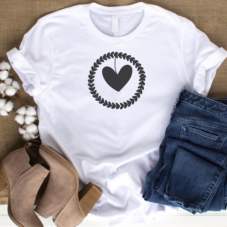 May include: White t-shirt featuring a black heart design within a laurel wreath. The shirt is paired with blue jeans and tan ankle boots. The design is simple and elegant, suitable for casual wear.