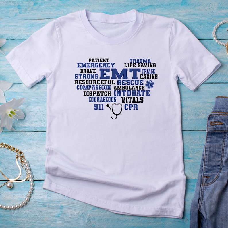 May include: White t-shirt with a blue graphic design that says "EMT" surrounded by words describing the job, such as "patient", "emergency", "trauma", "life saving", "strong", "resourceful", "rescue", "caring", "compassion", "ambulance", "dispatch", "intubate", "courageous", "vitals", "911", and "CPR".