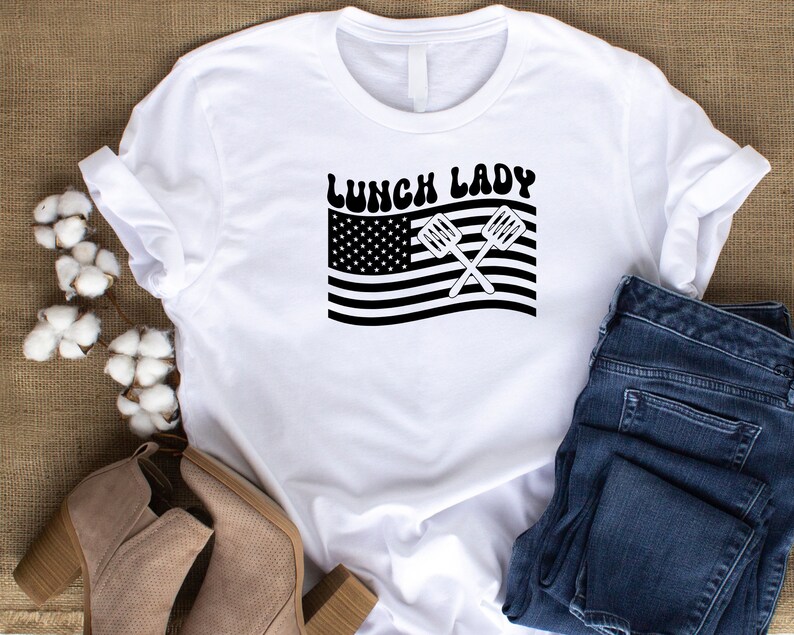 May include: White t-shirt with the text "Lunch Lady" in black letters with a retro font.  The text is above a black and white image of an American flag with two crossed spatulas.