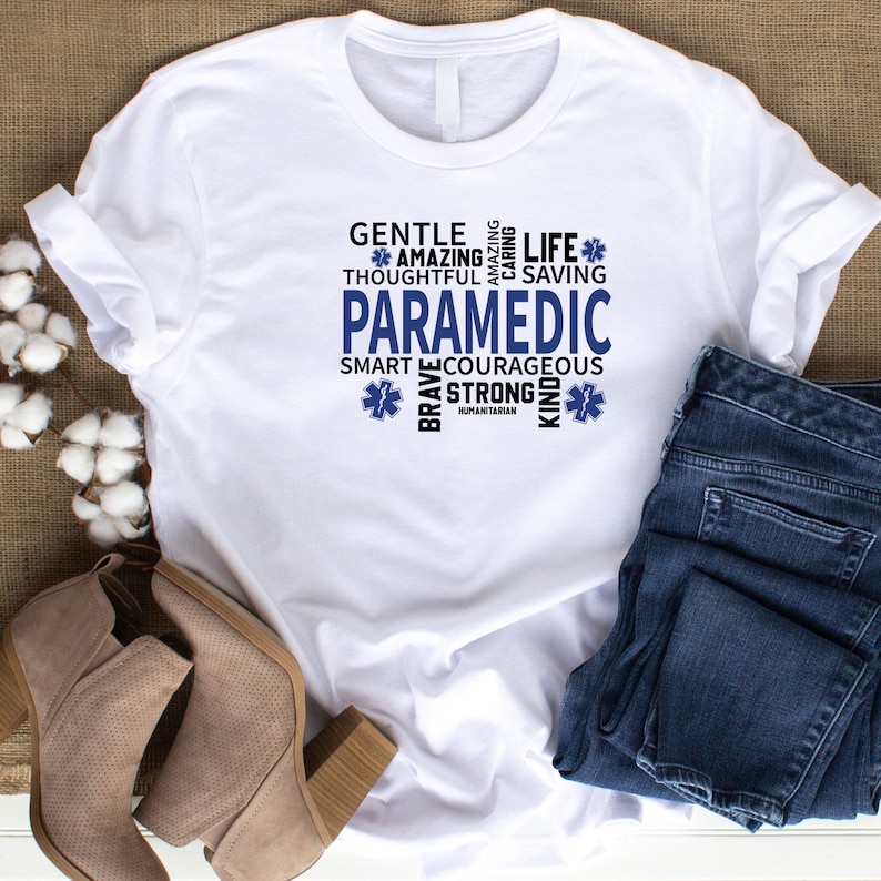 May include: White t-shirt with a blue and white graphic design that says "Paramedic" surrounded by words describing the qualities of a paramedic, such as "gentle", "amazing", "brave", "strong", "kind", and "life saving".