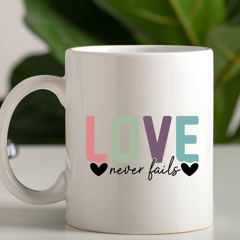 May include: White ceramic mug with a colorful "LOVE never fails" design. The letters are in pink, green, purple, and blue. There are two black hearts on either side of the text.