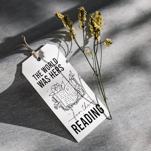May include: White gift tag with black text that reads "The world was hers for the reading" and a black and white illustration of an open book with flowers.