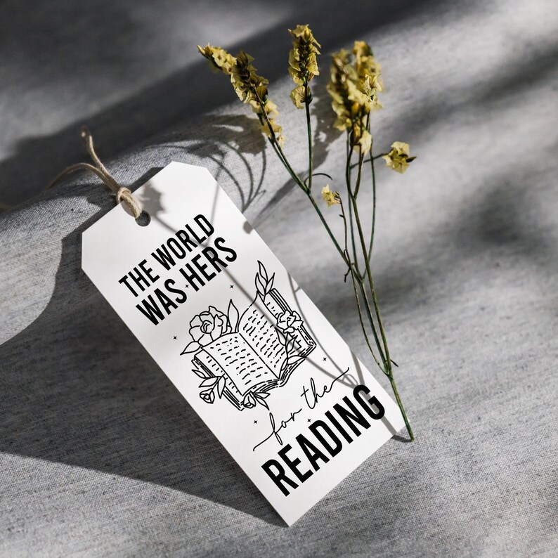 May include: White gift tag with black text that reads "The world was hers for the reading" and a black and white illustration of an open book with flowers.