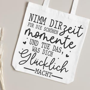 May include: White tote bag with black text that reads "Nimm dir Zeit f&uuml;r die sch&ouml;nen Momente und tue das, was dich gl&uuml;cklich macht." The bag has two handles and is made of a light-colored fabric.