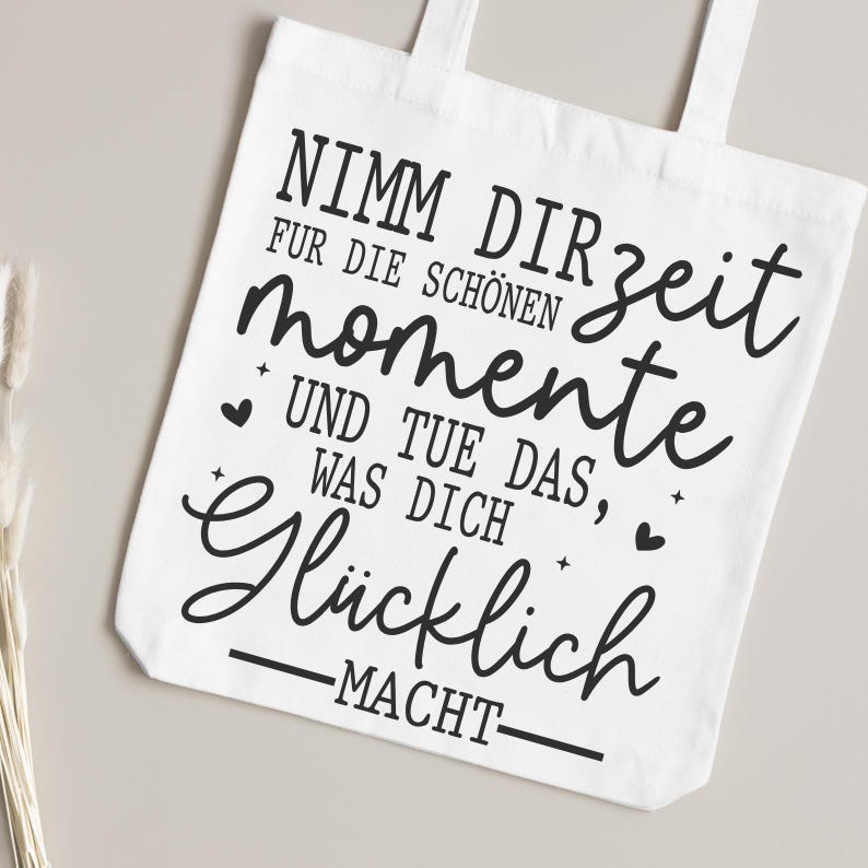 May include: White tote bag with black text that reads "Nimm dir Zeit f&uuml;r die sch&ouml;nen Momente und tue das, was dich gl&uuml;cklich macht." The bag has two handles and is made of a light-colored fabric.