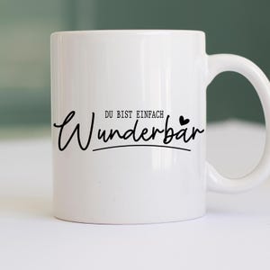May include: White ceramic mug with a black script that reads "Wunderb&auml;r" with the words "DU BIST EINFACH" above it. The mug has a curved handle and is set against a blurred background.