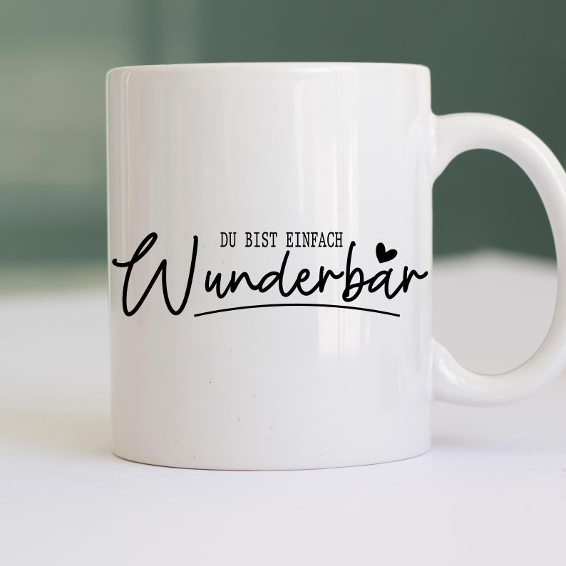May include: White ceramic mug with a black script that reads "Wunderb&auml;r" with the words "DU BIST EINFACH" above it. The mug has a curved handle and is set against a blurred background.