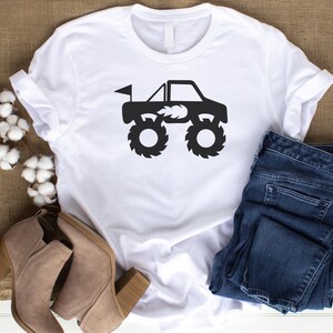 May include: White t-shirt with a black silhouette of a monster truck with flames on the side. The truck has a flag on the back and large tires.